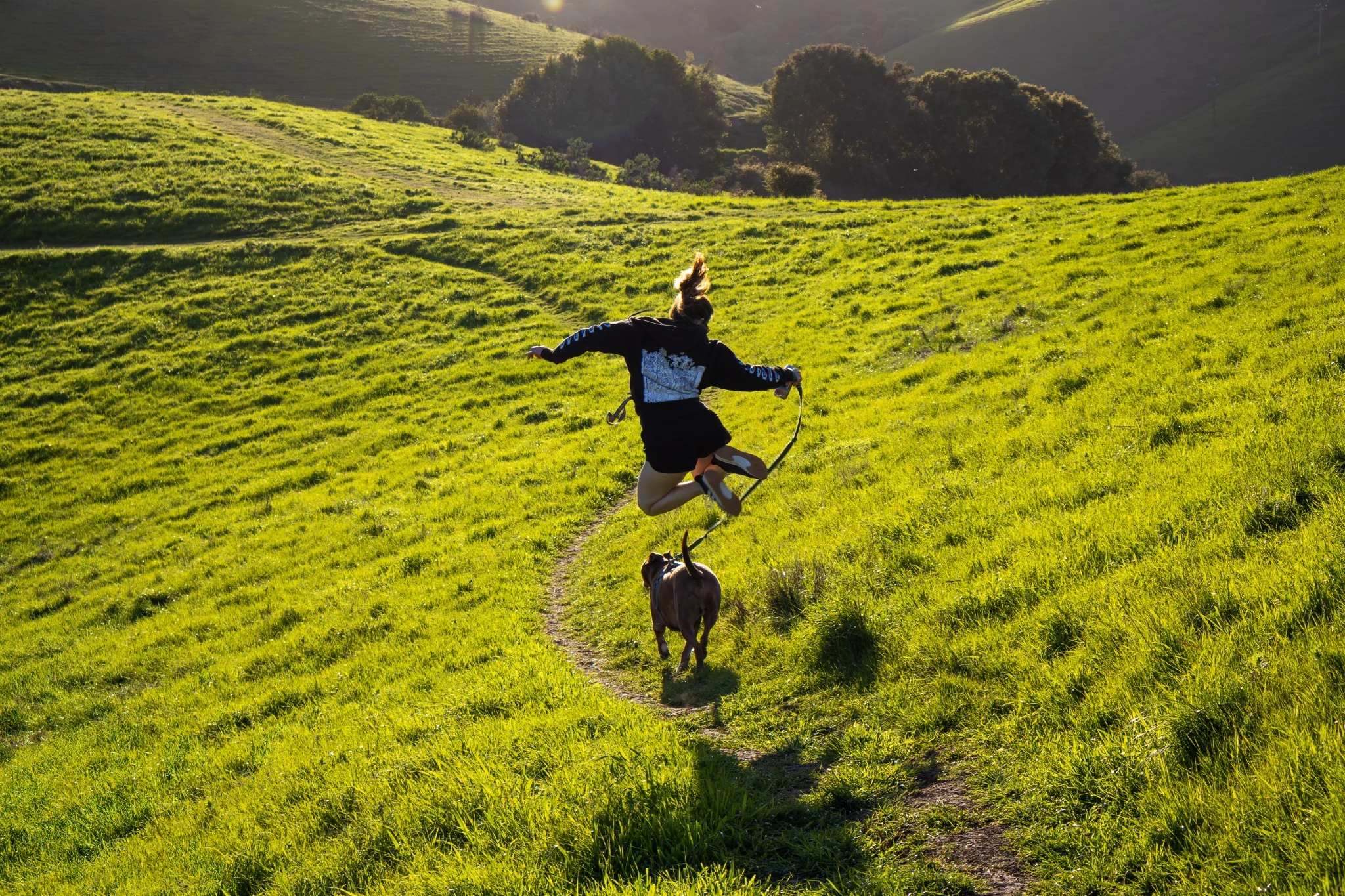 A person joyfully jumping on a sunlit green trail with a dog