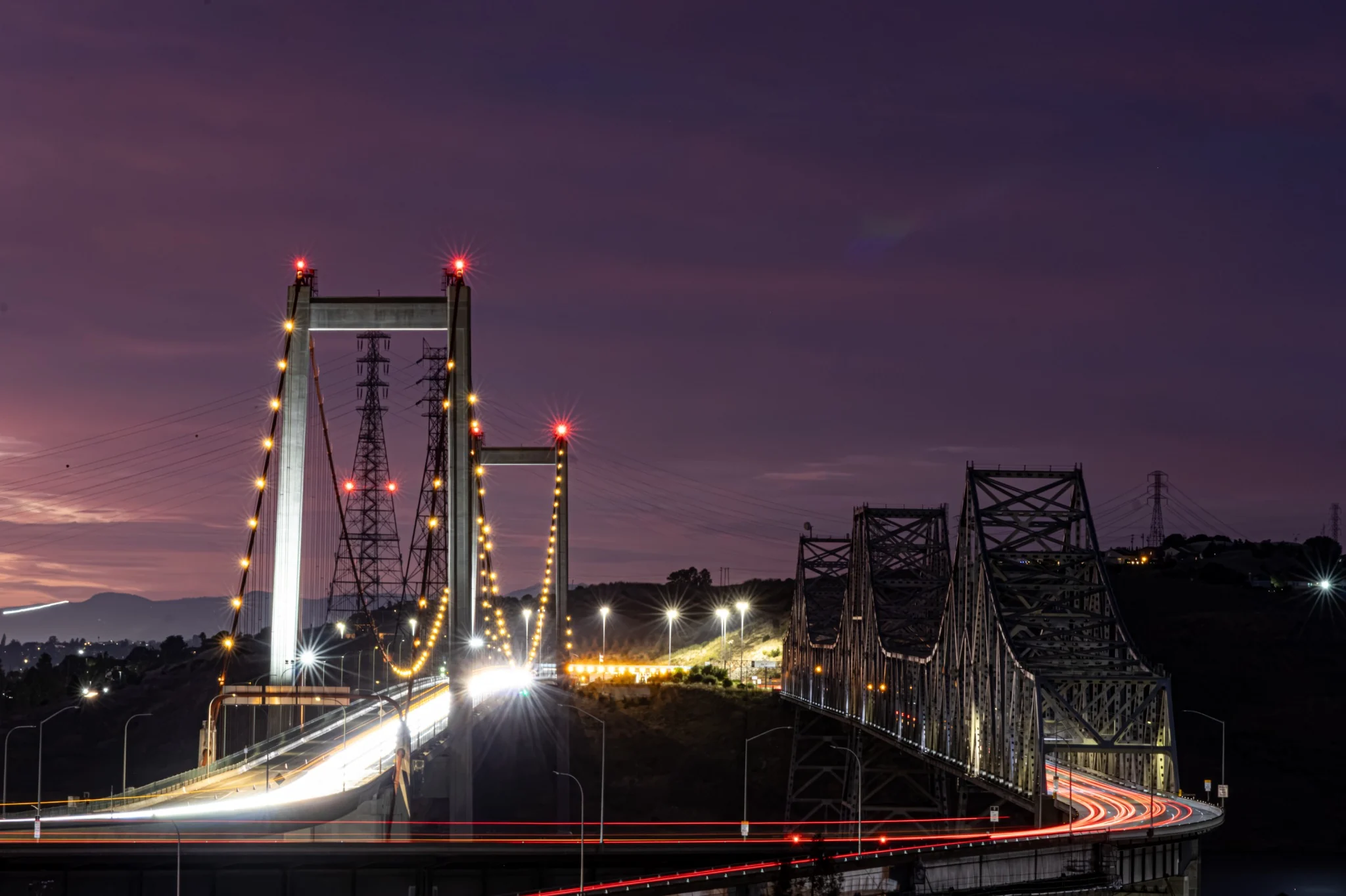 Long-exposure traffic light trails crossing the Carquinez Bridge beneath a purple evening sky